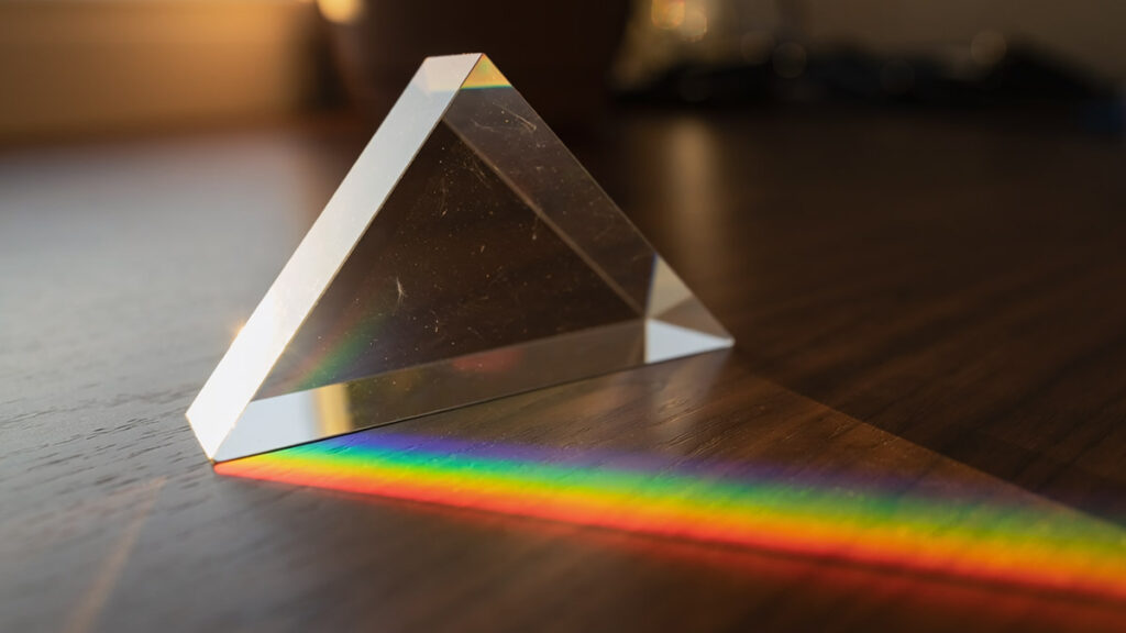 Ein Glasprisma auf einem Holztisch spaltet einfallendes Sonnenlicht in ein farbiges Regenbogenspektrum — veranschaulicht die Dispersion des Lichts (KI-generiert)