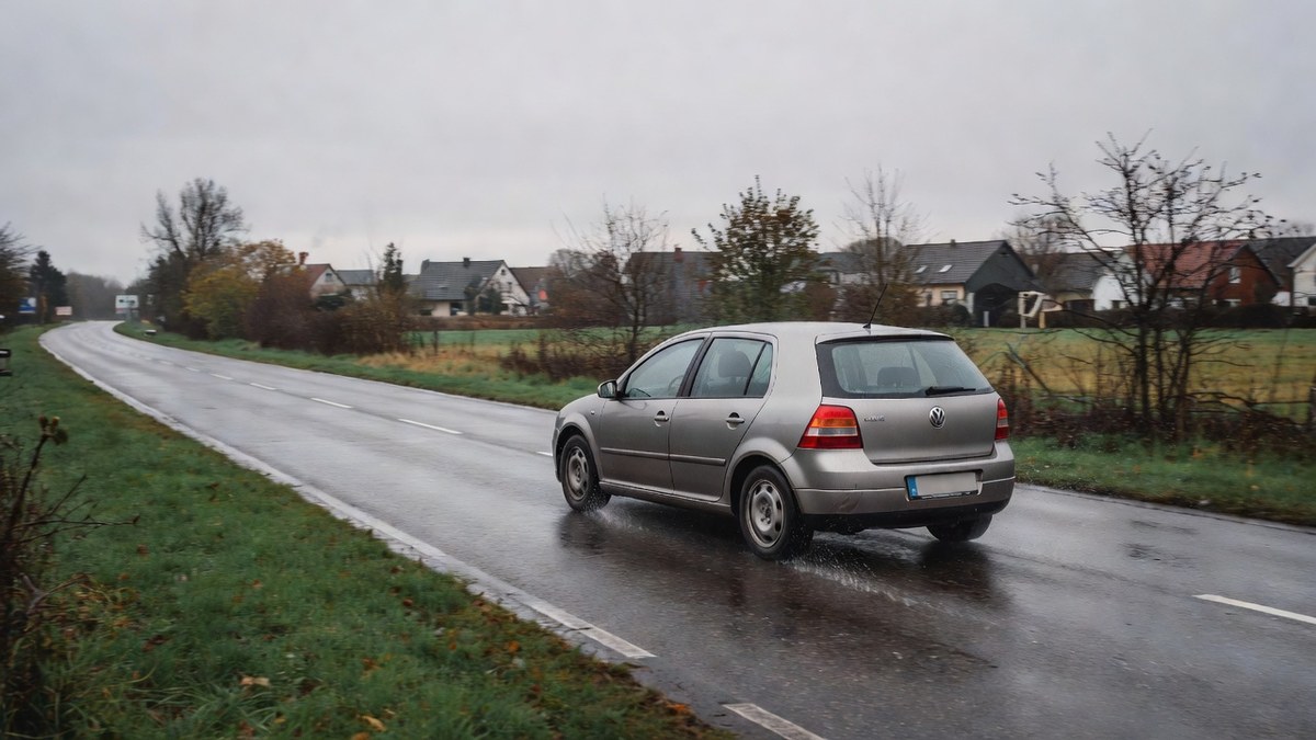 Ein kompaktes Auto fährt auf einer nassen Landstraße bei bedecktem Himmel, sinnbildlich für den Arbeitsweg und die Fahrtkosten in der Steuererklärung (KI-generiert)